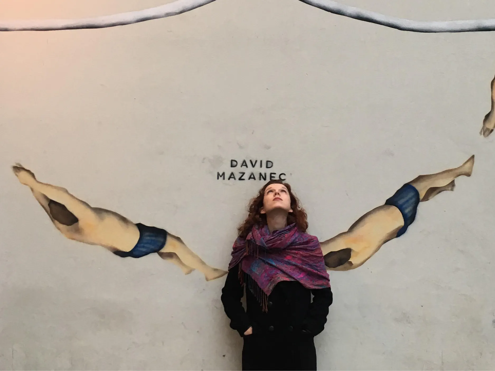 Mlada ženska stoji pred steno z grafiti plavalcev, za njeno glavo podpis avtorja David Mazanec